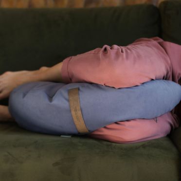 Osoba w różowych spodniach leży na ciemnozielonej kanapie, między kolanami ma poduszkę do medytacji i relaksacj w kolorze zgaszonym niebieskim.